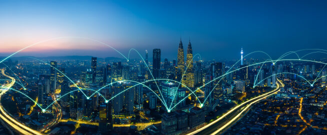 City skyline at dusk with rays of light representing network connectivity flashing in the sky from building to building.